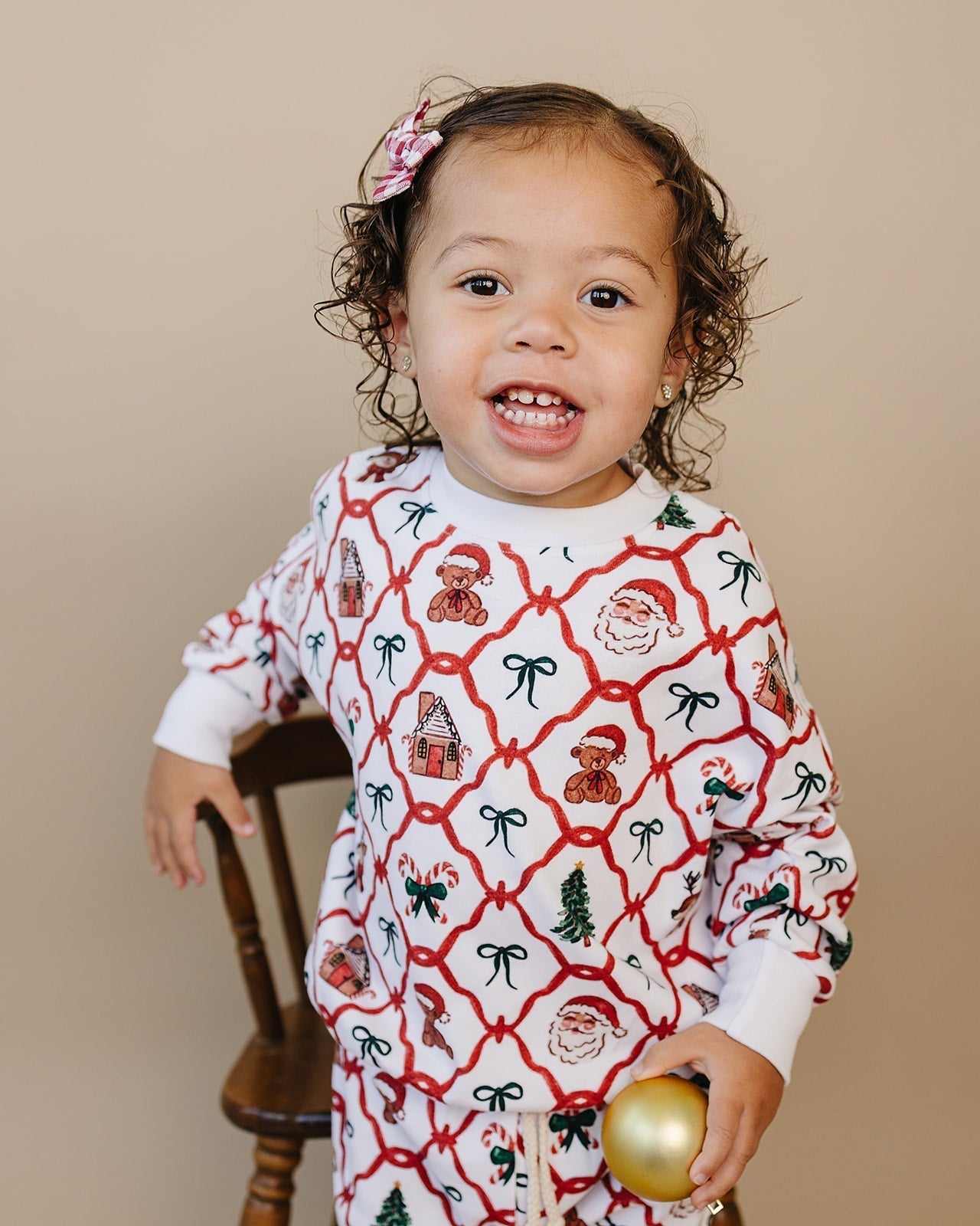 Smiling toddler in Christmas pajamas holding gold ornament on wooden chair